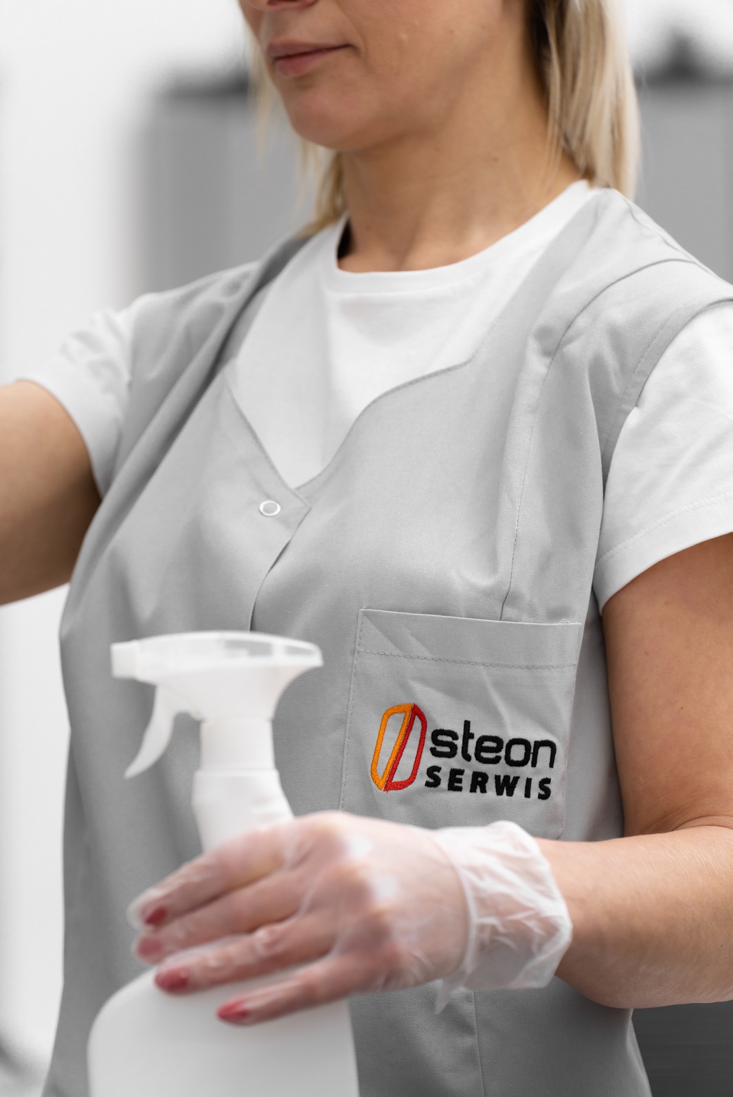 Pracownica w szarym uniformie z logo firmy 'Osteon Serwis' trzyma biały spryskiwacz, dłoń w przezroczystej rękawiczce, przygotowana do pracy.