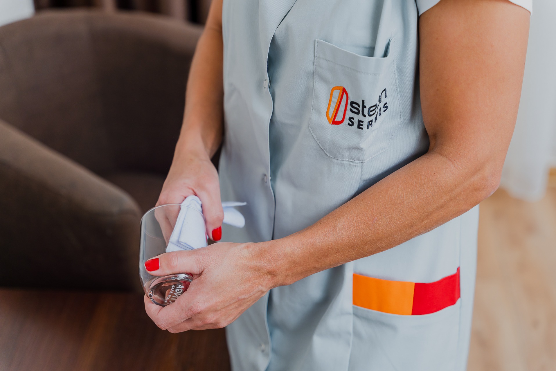 Kobieta w uniformie z logo firmy czyści szklankę ściereczką na tle brązowego fotela.