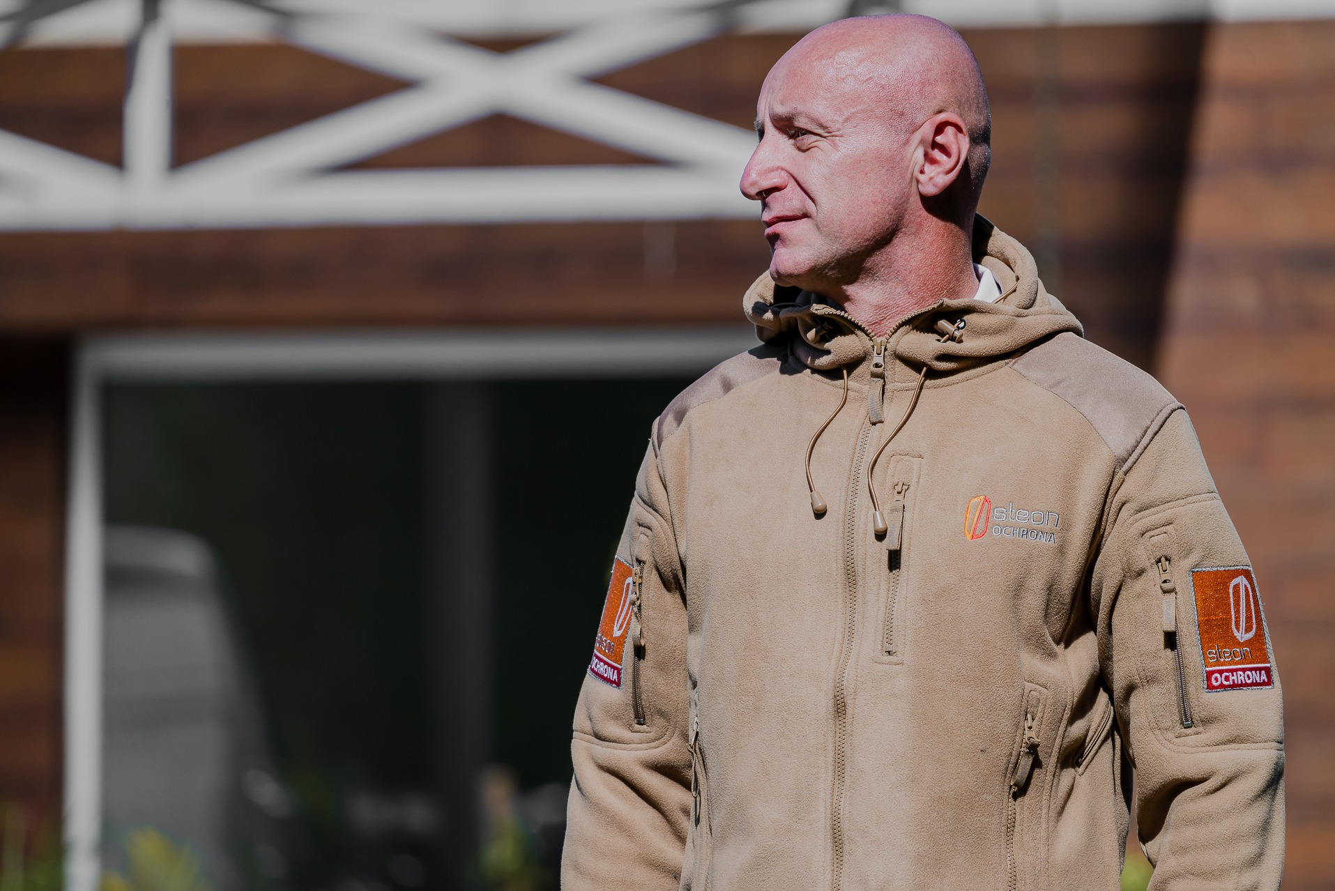 Mężczyzna w beżowej bluzie z logo firmy ochroniarskiej, patrzy w lewo, na tle domu z brązową elewacją.