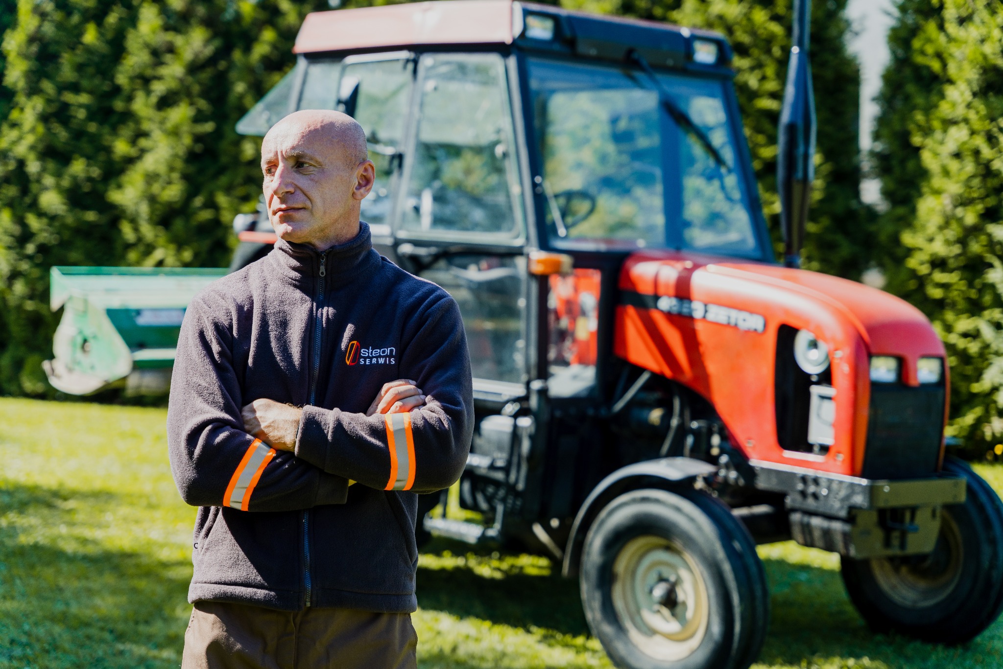 Mężczyzna z założonymi rękami w polarze z logo firmy na tle czerwonego traktora na trawniku w słoneczny dzień.