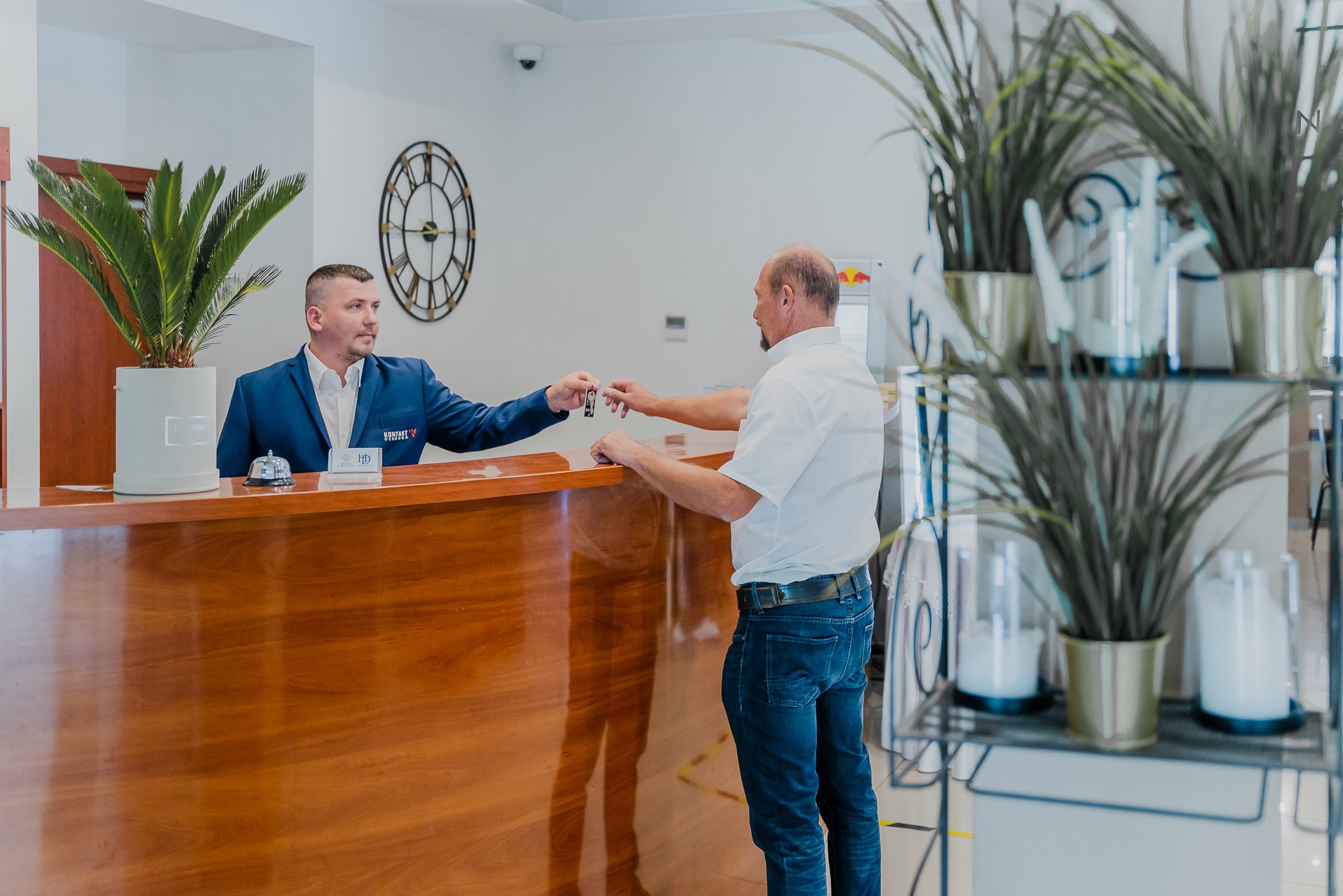 Recepcjonista w Niepołomicach wręcza klucze gościowi przy drewnianej ladzie z roślinami i zegarem na ścianie.