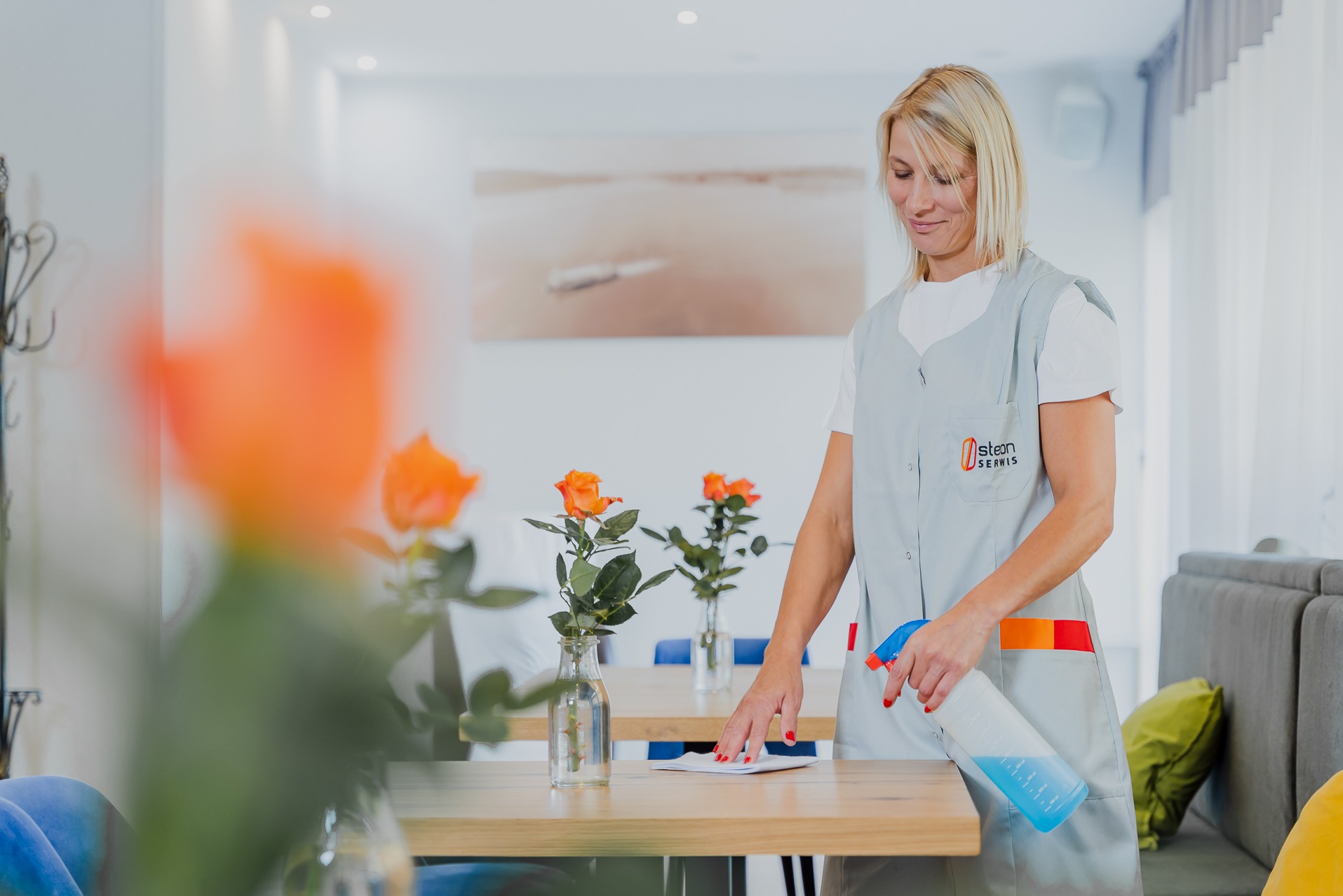 Pracownica w szarym uniformie z logo firmy spryskuje płynem czyszczącym blat stołu z bukietami pomarańczowych róż w wazonach, w tle jasne wnętrze biura z obrazem.