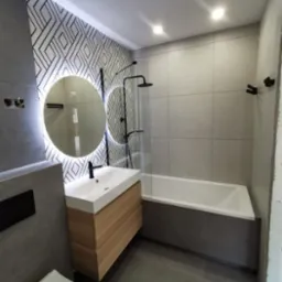 Nowoczesna łazienka w szarościach z akcentem drewna: okrągłe lustro LED, geometryczny wzór na ścianie, minimalistyczna armatura w czerni, wanna z szarą obudową.