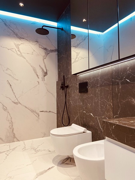 Elegancka łazienka z marmurowymi płytkami, podwieszaną toaletą i bidetem. Czarna deszczownica i oświetlenie LED dodają nowoczesnego charakteru.
