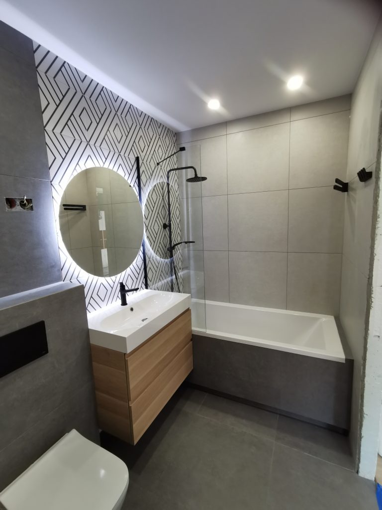 Nowoczesna łazienka w szarościach z akcentem drewna: okrągłe lustro LED, geometryczny wzór na ścianie, minimalistyczna armatura w czerni, wanna z szarą obudową.