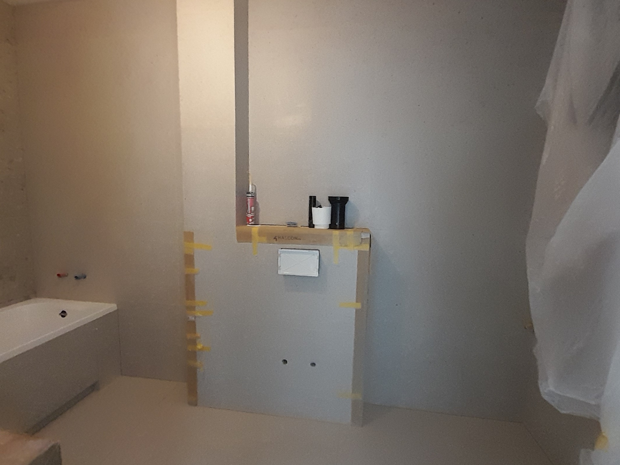 Wnętrze łazienki w trakcie prac: zabudowa z płyty gipsowej, częściowo zakryta folią, widoczna wanna. Przygotowania do wykończenia.