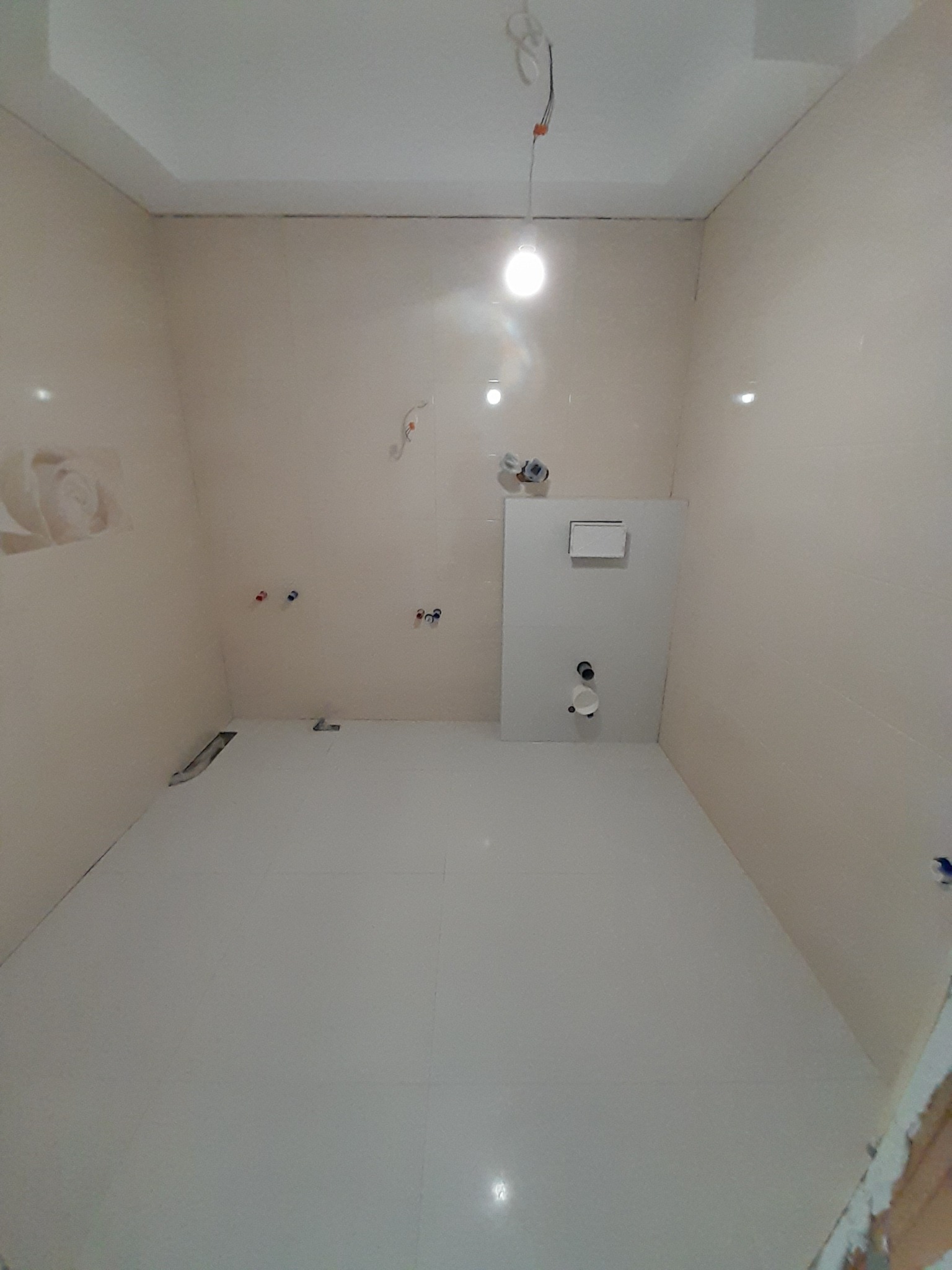 Wykończona łazienka z beżowymi ścianami i białymi płytkami podłogowymi, widoczne instalacje wodne i stelaż podtynkowy WC. Świeżo położone płytki.