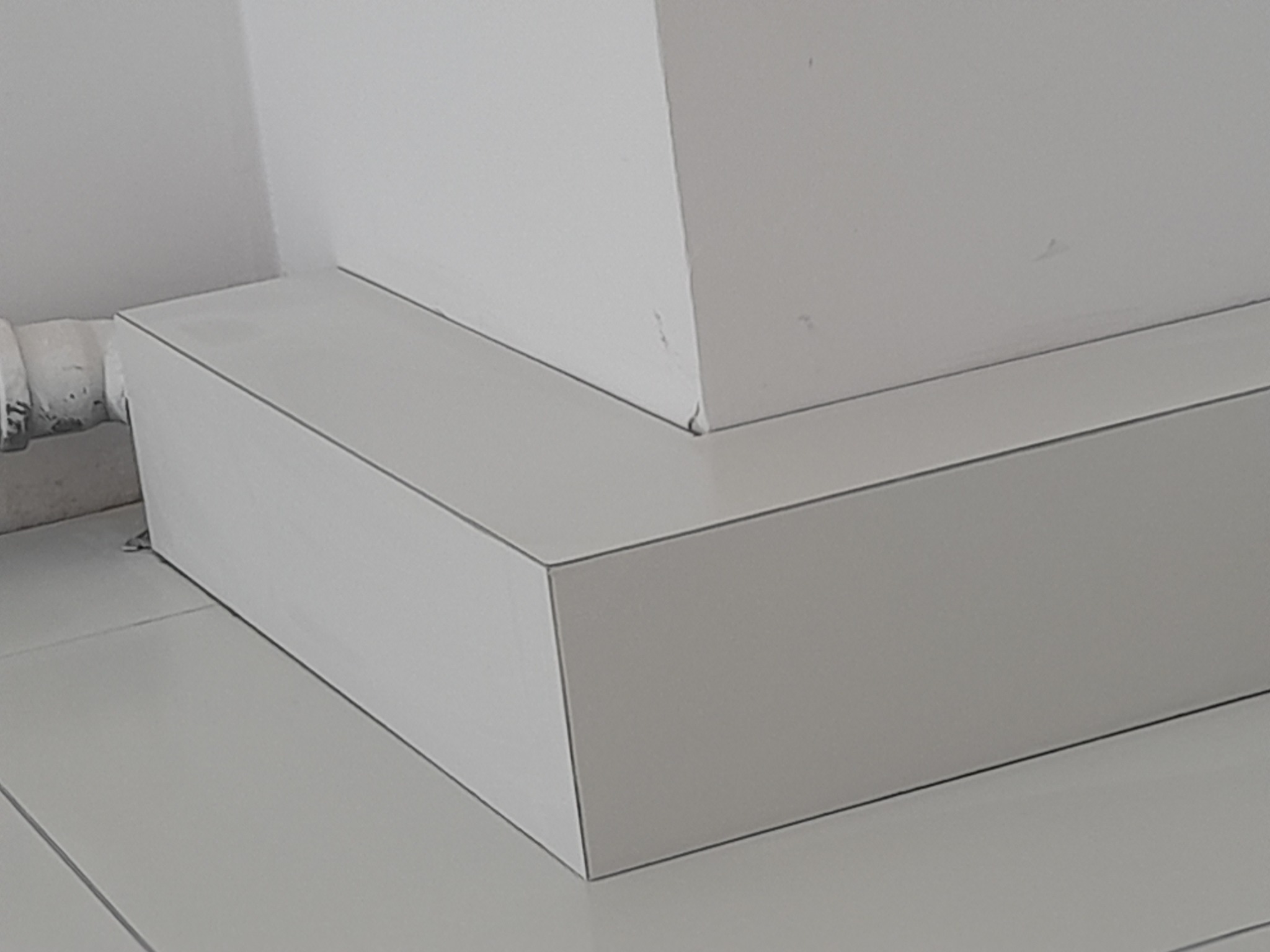 Białe, precyzyjnie wykonane obudowy rur i narożników w pomieszczeniu wykończonym białymi płytkami podłogowymi. Detal wykończenia wnętrza.