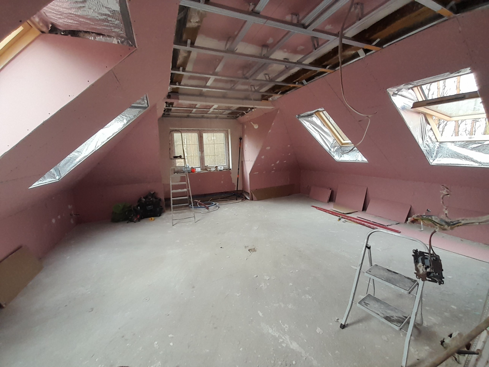 Poddasze w trakcie adaptacji: różowe płyty na skosach, metalowa konstrukcja sufitu, okna dachowe z folią. Widoczne drabiny i narzędzia.
