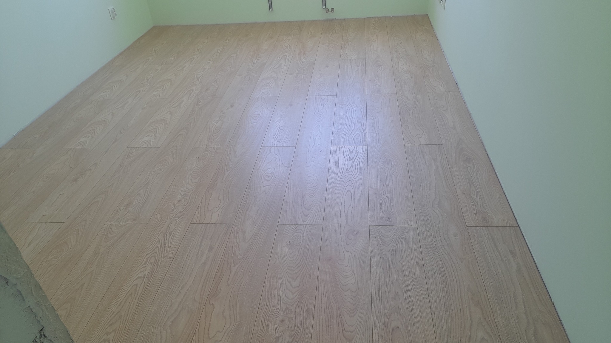 Ułożone panele podłogowe o jasnym, drewnopodobnym wzorze w pokoju z jasnozielonymi ścianami. Widoczne fragmenty ścian i listew przypodłogowych.