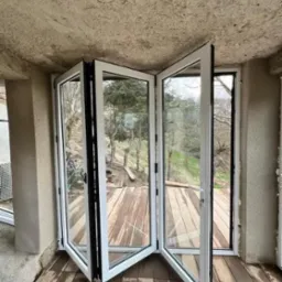 Otwarta, składana szklana ściana z białymi ramami, wychodząca na drewniany taras z widokiem na zieleń. Specyficzny surowy sufit i podłoga z desek.