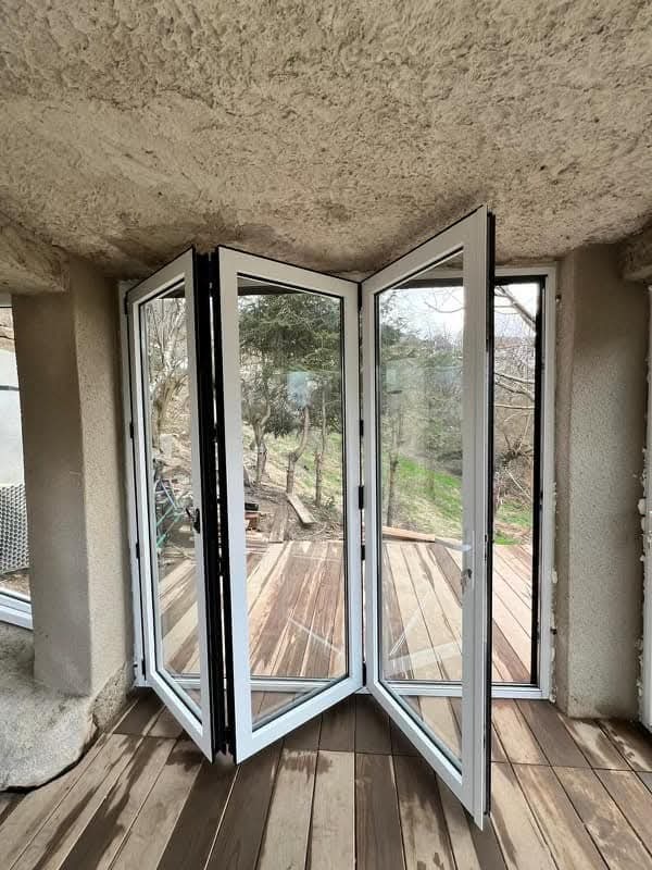 Otwarta, składana szklana ściana z białymi ramami, wychodząca na drewniany taras z widokiem na zieleń. Specyficzny surowy sufit i podłoga z desek.