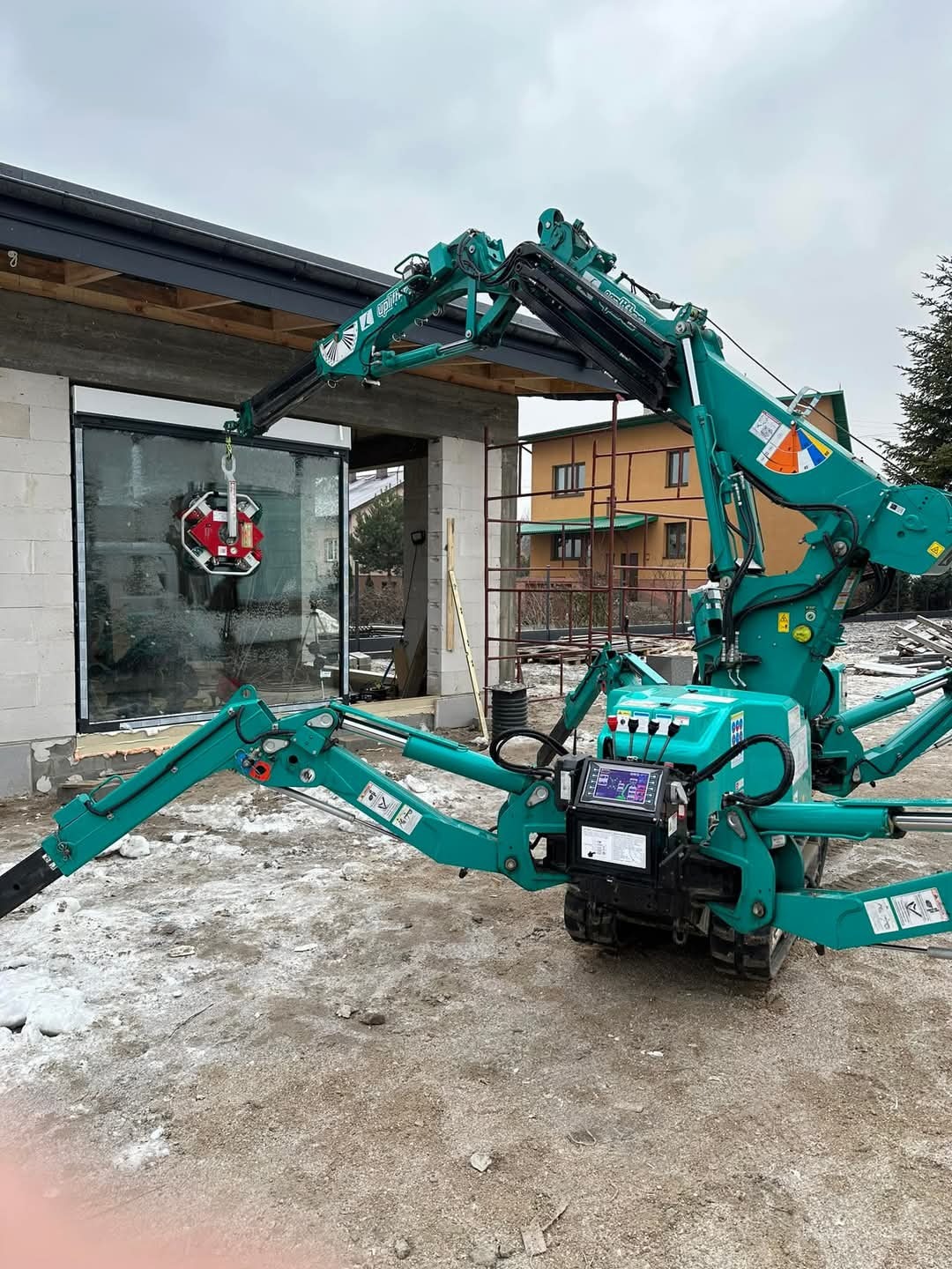 Montaż okna w surowym budynku za pomocą specjalistycznego dźwigu ssącego. Widoczny fragment domu w budowie i otoczenie.