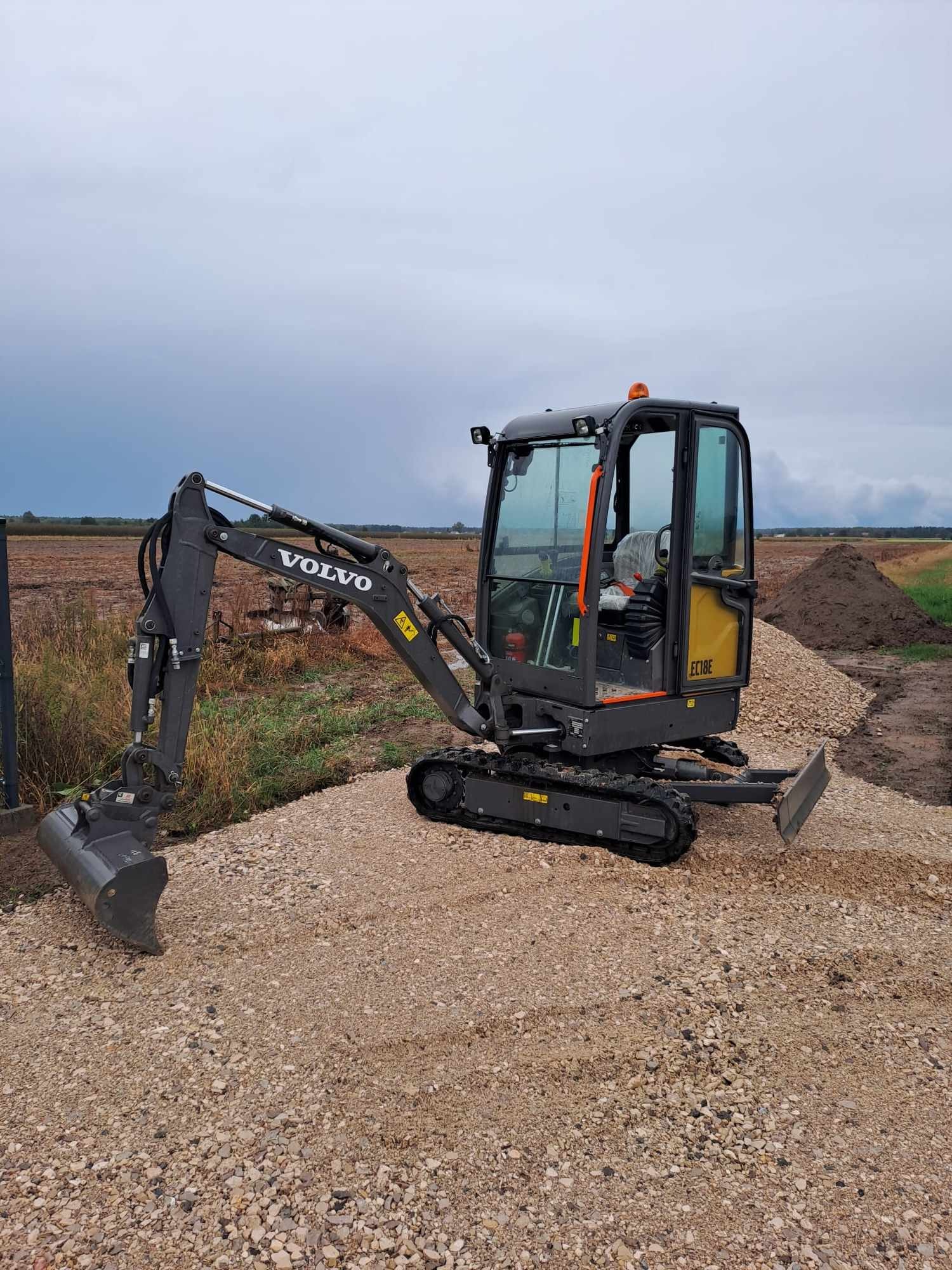 Mała koparka Volvo EC18E stoi na warstwie żwiru, w tle pole i pochmurne niebo, widoczny mały pryzmy ziemi.