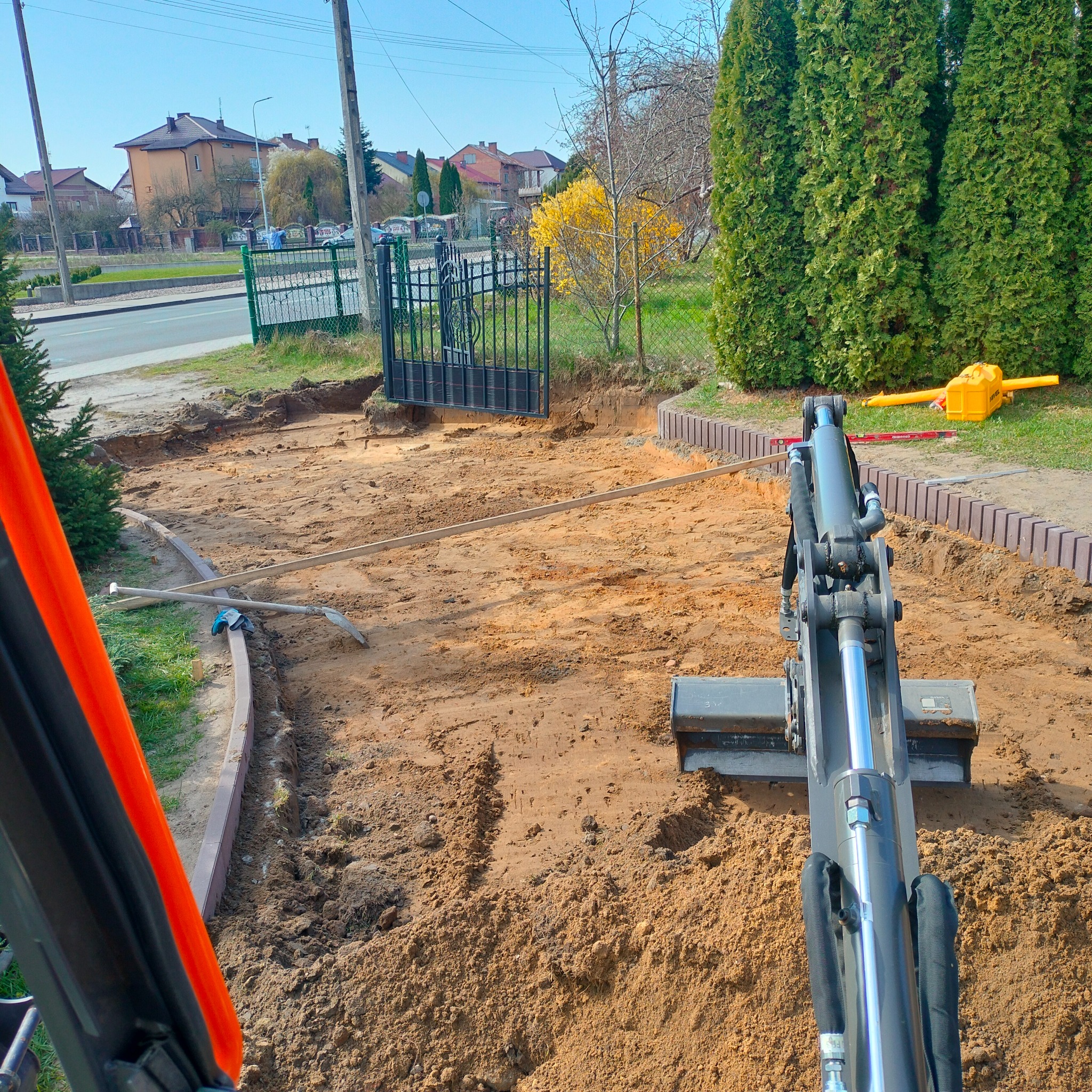 Mini koparka w trakcie prac ziemnych na posesji, widoczne wykopane fundamenty pod ogrodzenie i przygotowany teren pod trawnik, w tle brama wjazdowa i zabudowa jednorodzinna.