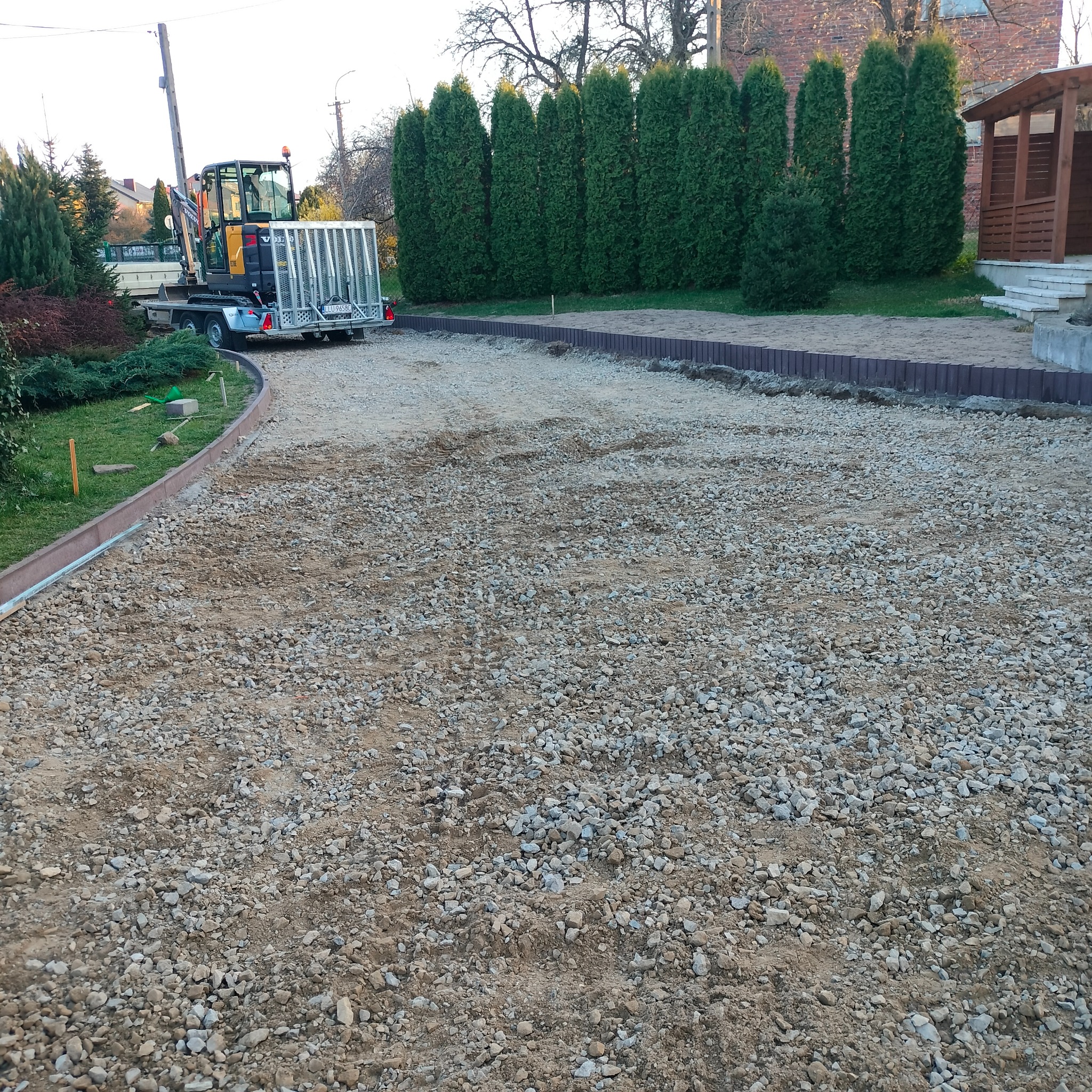 Przygotowany teren pod budowę z ułożoną obrzeżami, widoczny mini-koparka na przyczepie i rząd tuj.