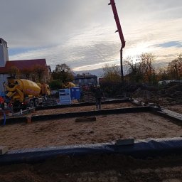 MBUD Usługi Budowlane IHOR SHEVCHUK - Przygotowanie terenu pod wylewkę fundamentów, widoczny szalunek, zbrojenie i betonowóz z pompą na tle kościoła pod pochmurnym niebem.