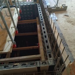 MBUD Usługi Budowlane IHOR SHEVCHUK - Szczegółowy widok szalunku zbrojeniowego fundamentu z wystającymi prętami zbrojeniowymi, drewnianymi wzmocnieniami i fragmentem muru z cegły w tle.