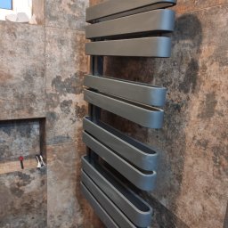 MBUD Usługi Budowlane IHOR SHEVCHUK - Szary, nowoczesny grzejnik drabinkowy zamontowany na ścianie z płytek imitujących kamień, widoczne akcesoria montażowe w niszy ściennej.