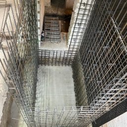 MBUD Usługi Budowlane IHOR SHEVCHUK - Zbrojenie fundamentu w wykopie, widoczne pręty stalowe tworzące szkielet, częściowo zalany beton, w tle schody prowadzące do piwnicy, pracownik w odblaskowej kamizelce w rogu kadru.