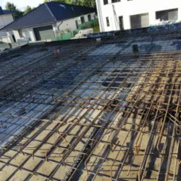 Zbrojenie płyty fundamentowej domu jednorodzinnego z widocznymi elementami dystansowymi z tworzywa sztucznego, przygotowane do zalania betonem, z domami w tle.