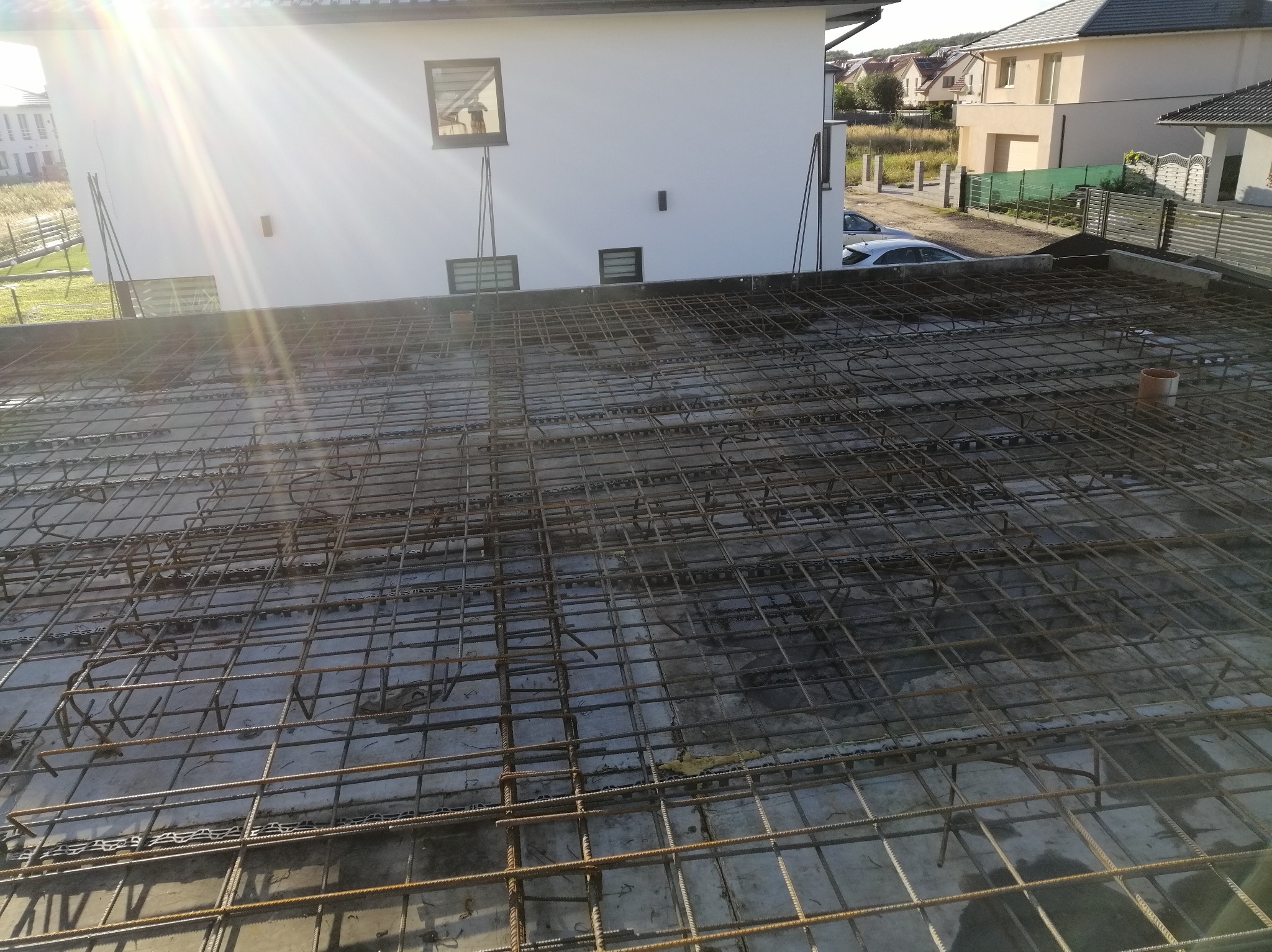 Zbrojenie płyty fundamentowej na wylewce betonowej, widok z góry na siatkę z prętów stalowych, w tle biały budynek w budowie i promienie słoneczne.