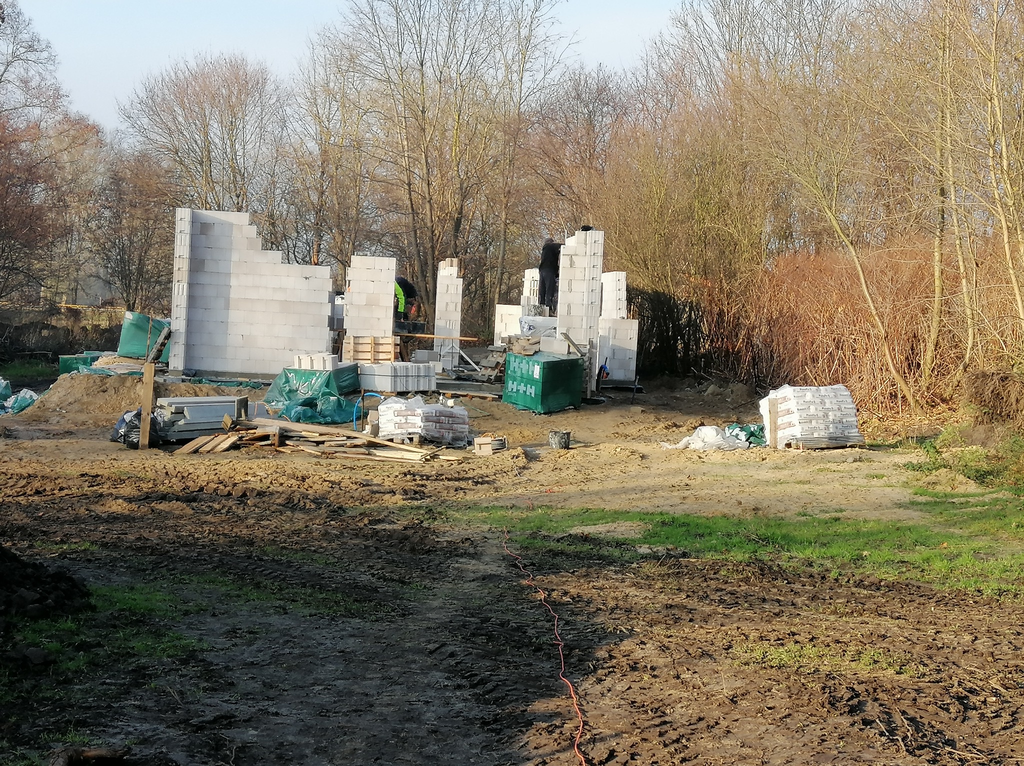 Wznoszenie ścian parteru z bloczków betonu komórkowego na placu budowy, widoczne narzędzia i materiały budowlane, w tle drzewa bez liści.