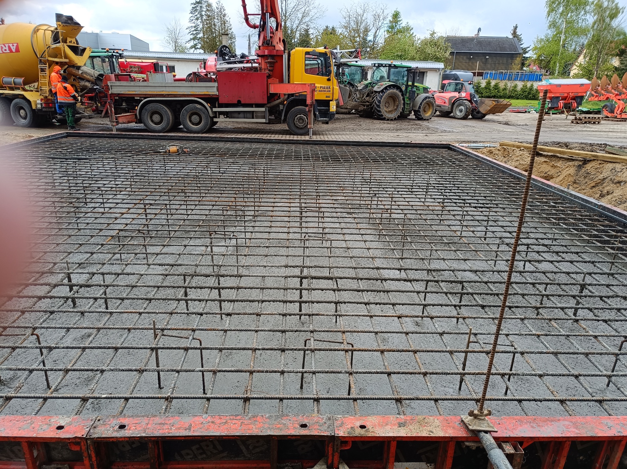 Zalewanie betonem zbrojonej konstrukcji szkieletowej fundamentu z widocznymi maszynami budowlanymi w tle.