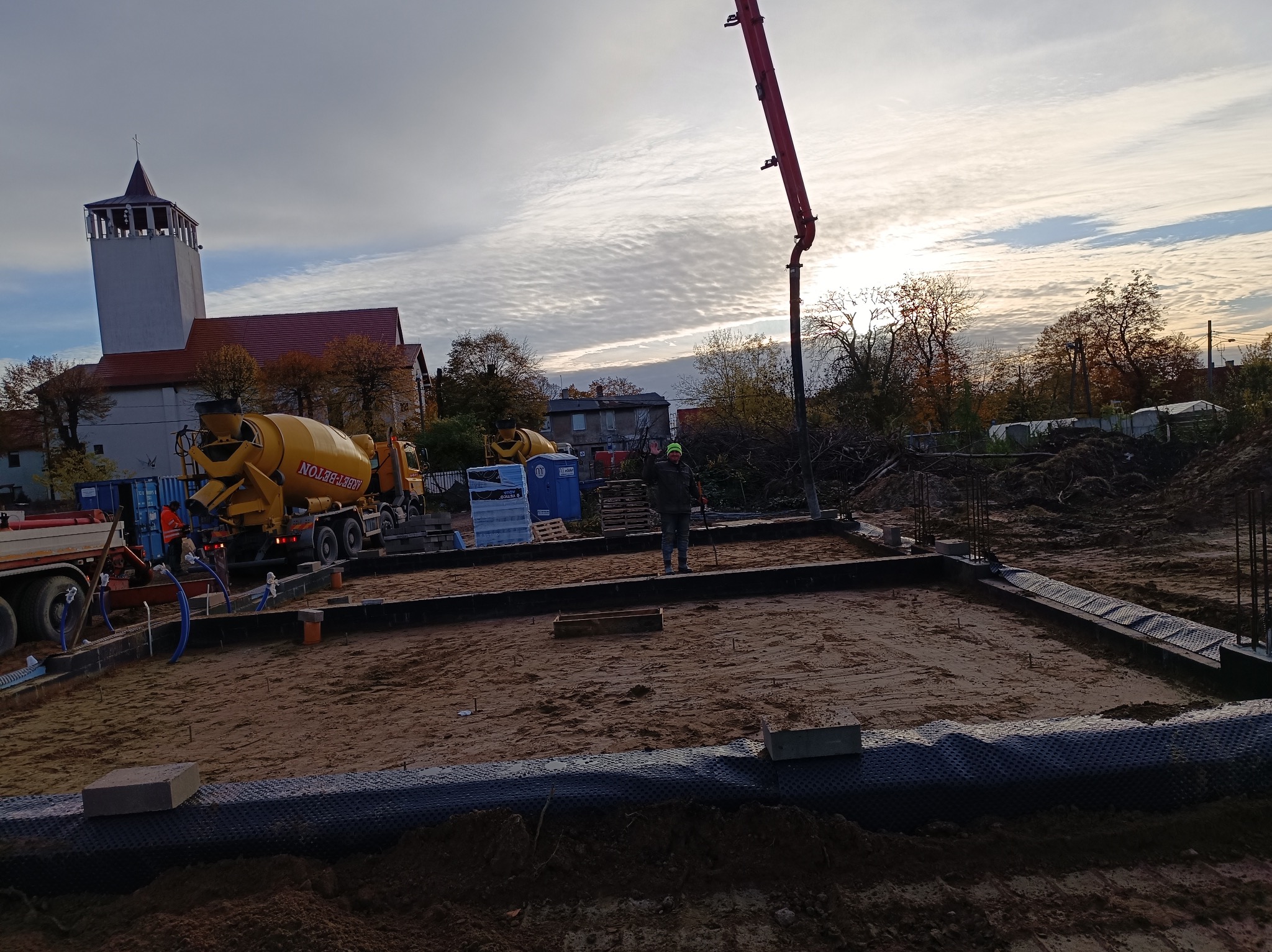 Przygotowanie terenu pod wylewkę fundamentów, widoczny szalunek, zbrojenie i betonowóz z pompą na tle kościoła pod pochmurnym niebem.