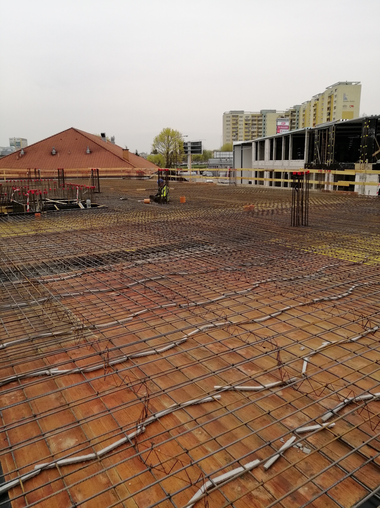 Zbrojenie stropu budynku w trakcie budowy, widoczne pręty zbrojeniowe, szalunki, betonowe dystanse oraz pracownik w oddali, w tle budynki mieszkalne.