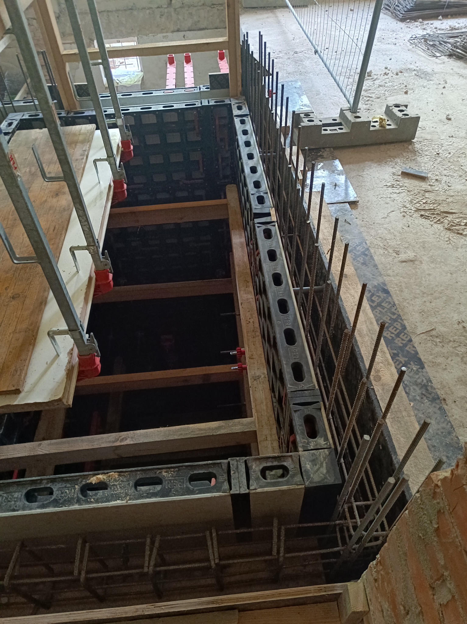 Szczegółowy widok szalunku zbrojeniowego fundamentu z wystającymi prętami zbrojeniowymi, drewnianymi wzmocnieniami i fragmentem muru z cegły w tle.