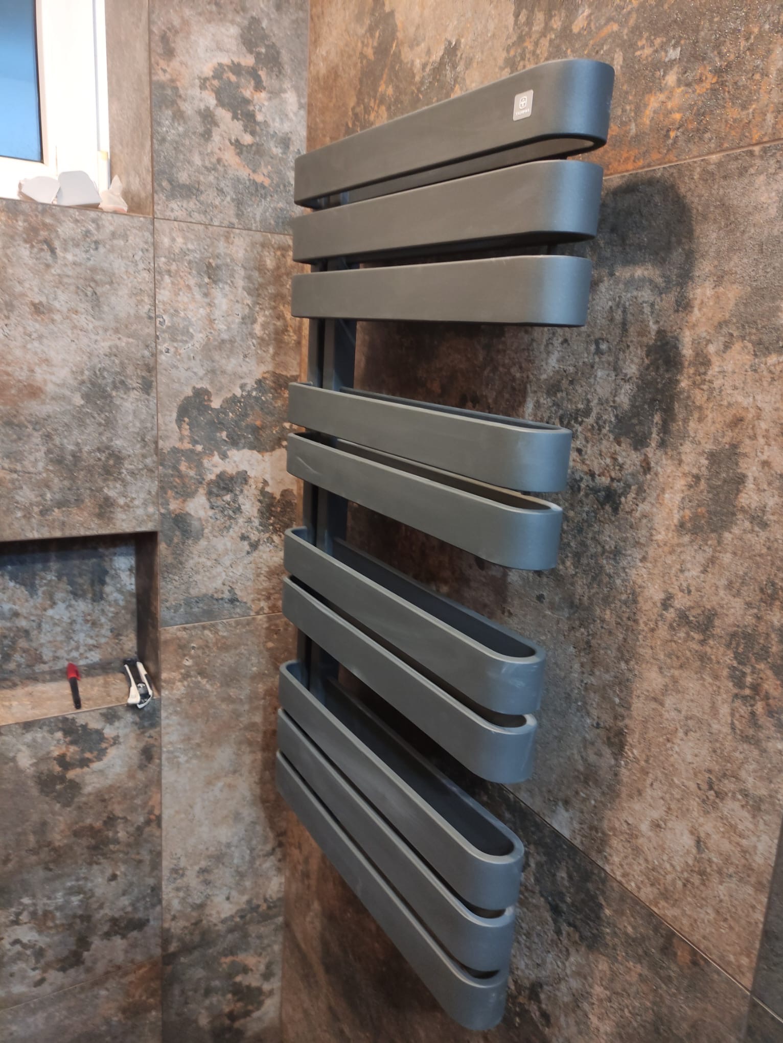 Szary, nowoczesny grzejnik drabinkowy zamontowany na ścianie z płytek imitujących kamień, widoczne akcesoria montażowe w niszy ściennej.