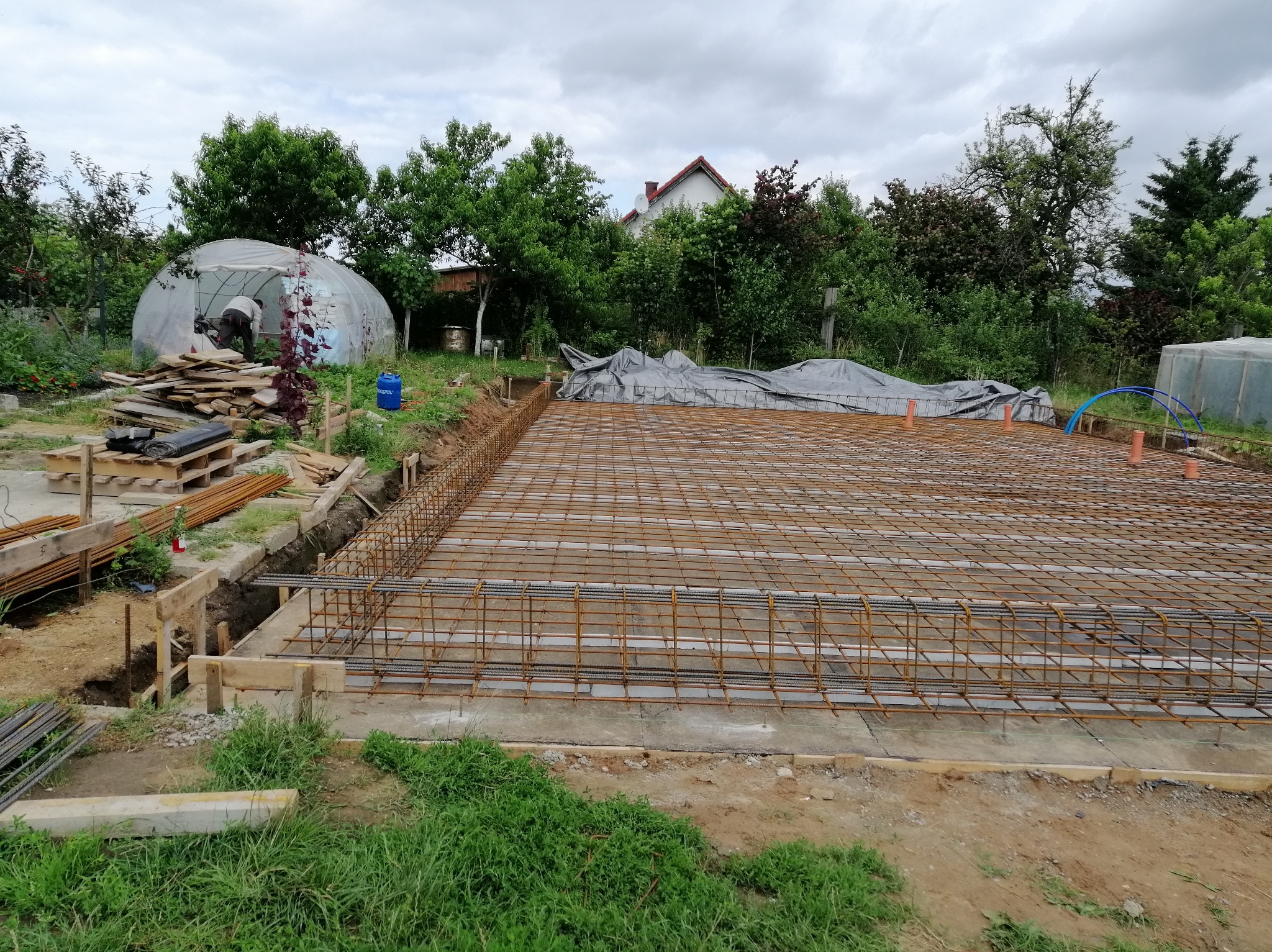 Zbrojenie fundamentu domu jednorodzinnego z widocznym szalunkiem drewnianym i folią pod zbrojeniem, w tle szklarnia i zieleń ogrodu pod pochmurnym niebem.
