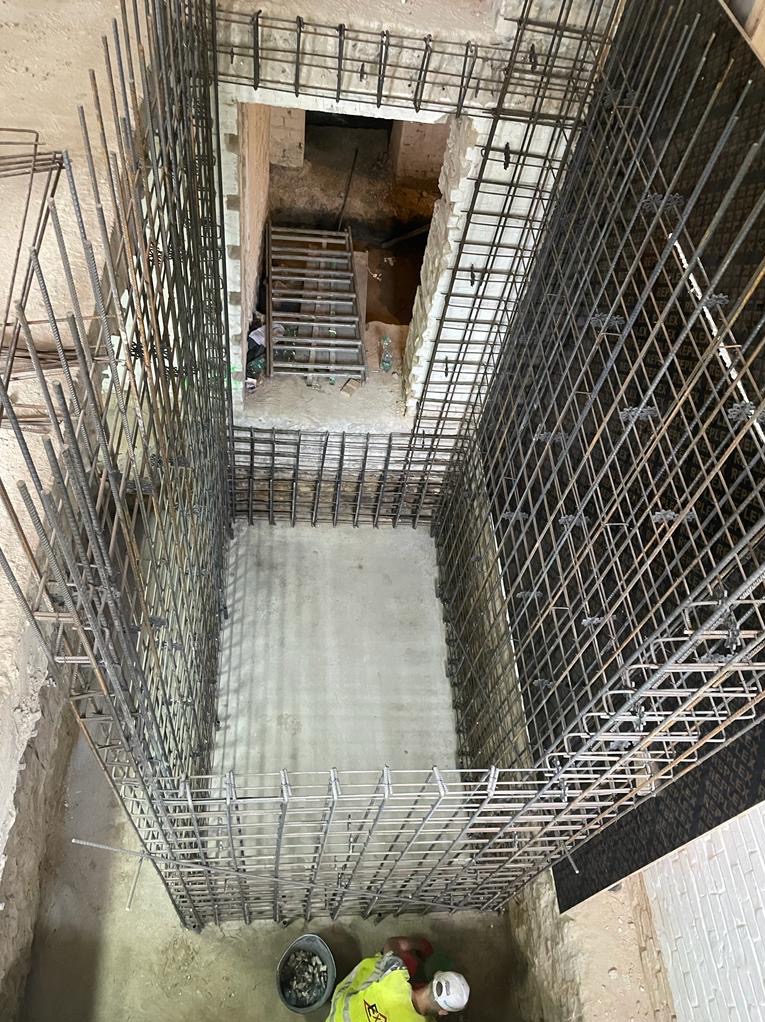 Zbrojenie fundamentu w wykopie, widoczne pręty stalowe tworzące szkielet, częściowo zalany beton, w tle schody prowadzące do piwnicy, pracownik w odblaskowej kamizelce w rogu kadru.