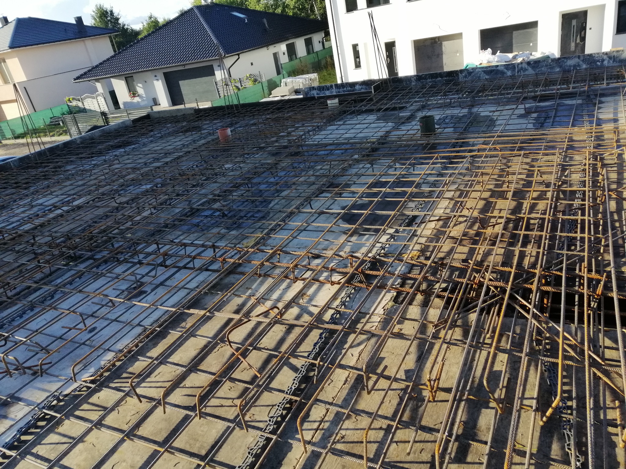 Zbrojenie płyty fundamentowej domu jednorodzinnego z widocznymi elementami dystansowymi z tworzywa sztucznego, przygotowane do zalania betonem, z domami w tle.