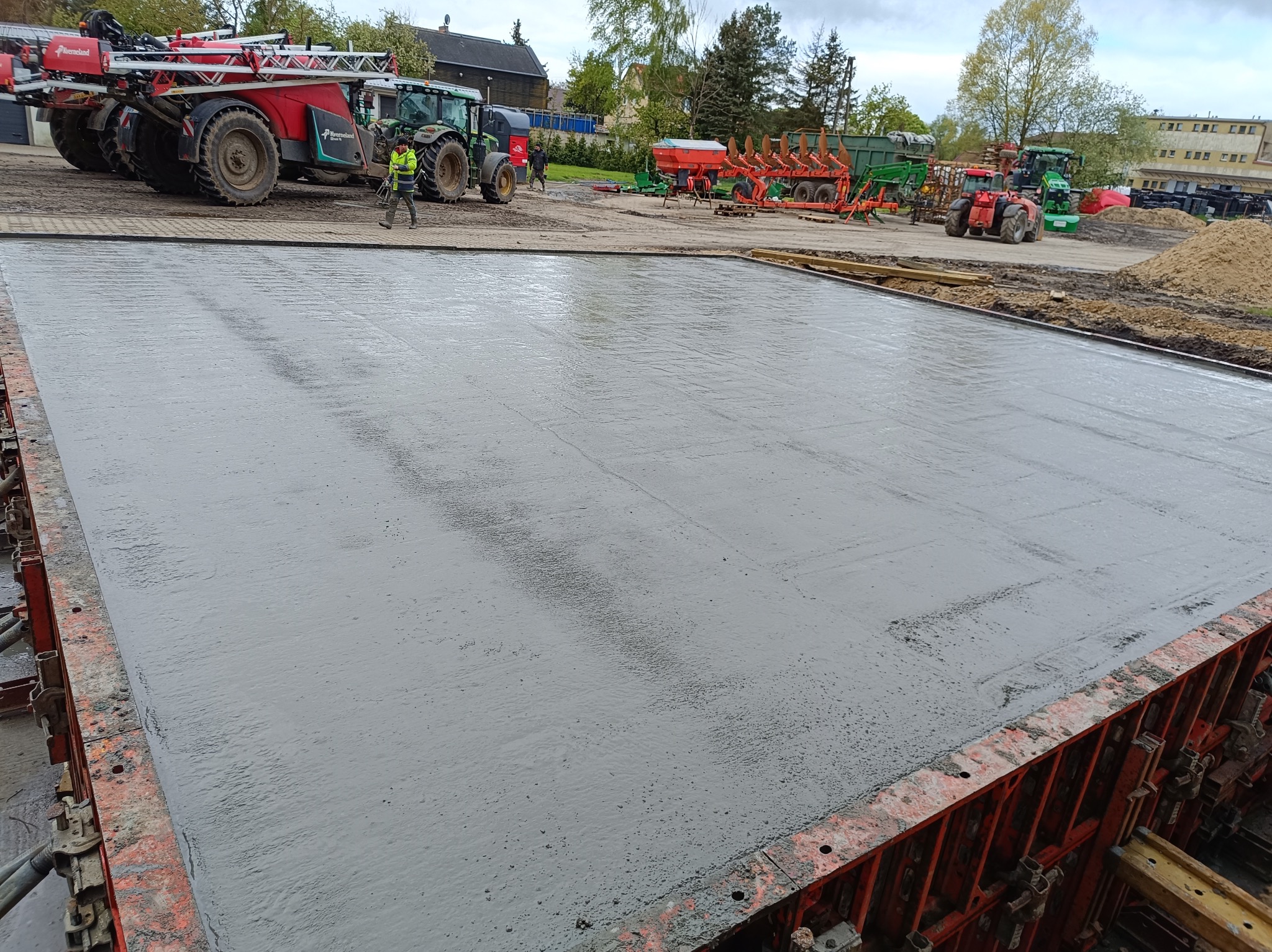 Świeżo wylany, mokry fundament z betonu w pomarańczowym szalunku, w tle maszyny rolnicze i budynki.