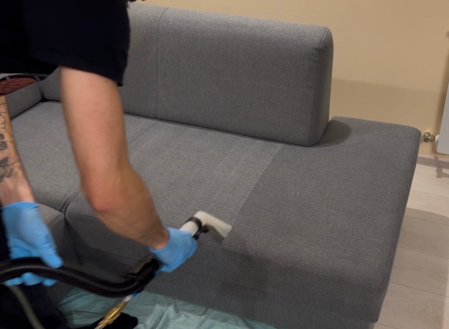 Czyszczenie narożnika z szarej tapicerki. Widoczna różnica w odcieniu po wypraniu fragmentu. Pracownik w niebieskich rękawiczkach używa ssawki do tapicerki.