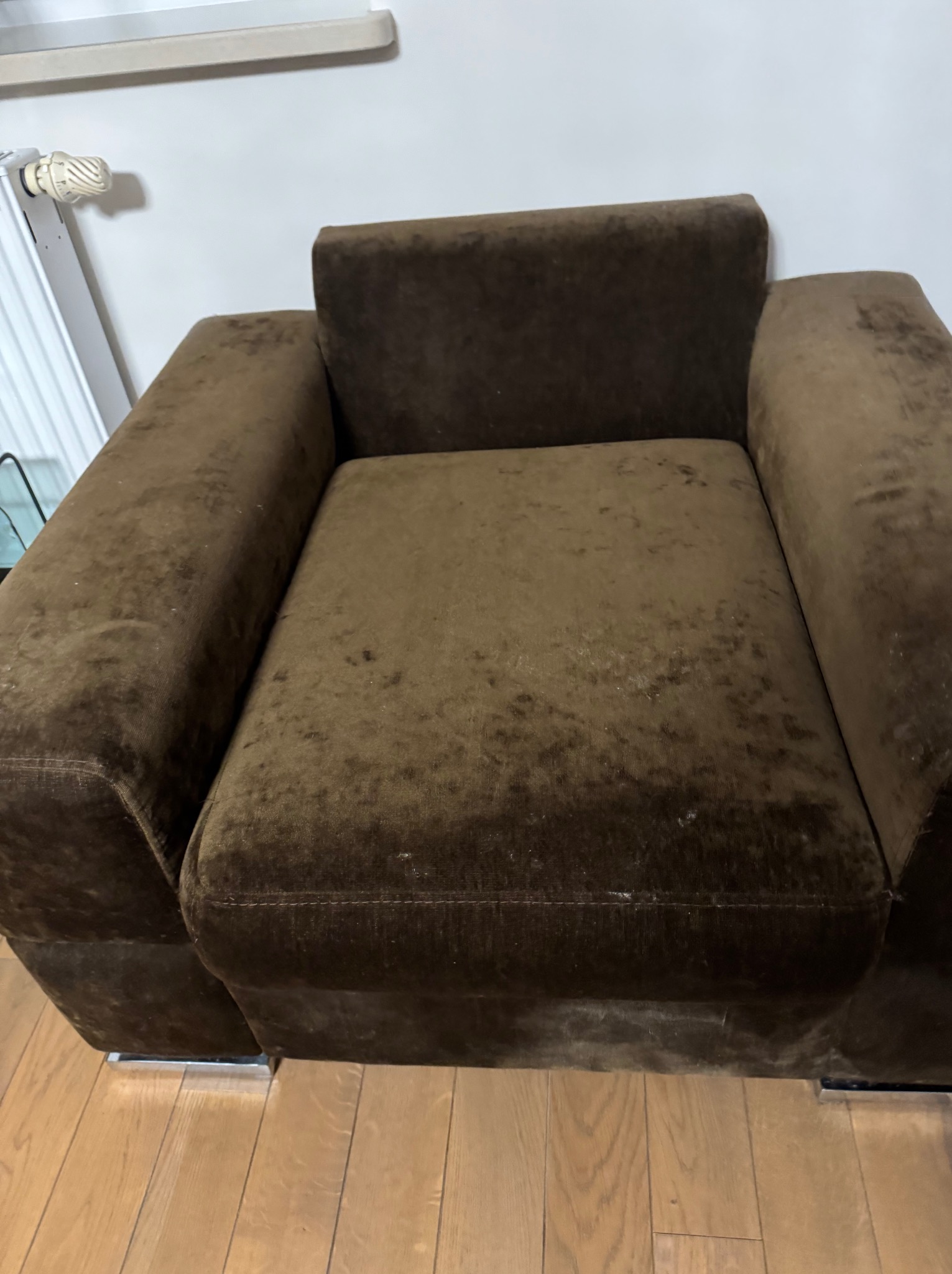 Brązowy fotel z welurową tapicerką, widoczne plamy i zabrudzenia na siedzisku i podłokietnikach, stojący na drewnianej podłodze, z grzejnikiem w tle.