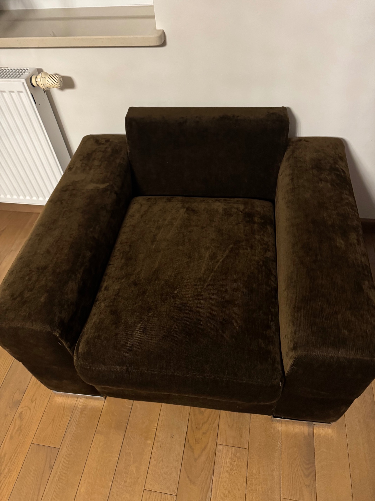 Ciemnobrązowy, welurowy fotel z podłokietnikami i niskim oparciem, stojący na drewnianej podłodze, z grzejnikiem w tle.