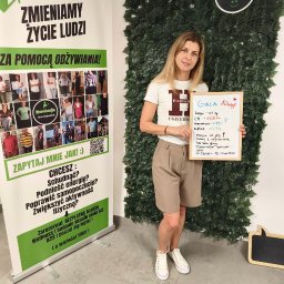 KLUB ZDROWEGO STYLU ŻYCIA MAGDALENA LESZCZYŃSKA - Uśmiechnięta kobieta w białej koszulce prezentuje na tablicy wyniki klienta (spadek wagi, wzrost energii) obok roll-upu 'Ewolucja Klub Odżywiania' i zielonej ściany z liści.
