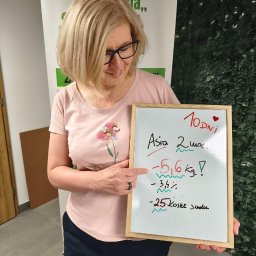 KLUB ZDROWEGO STYLU ŻYCIA MAGDALENA LESZCZYŃSKA - Uśmiechnięta kobieta w okularach, w różowej koszulce, wskazuje palcem na tablicę z odręcznymi wynikami utraty wagi: -5,6 kg i -25 kostek cukru, na tle zielonej ściany.