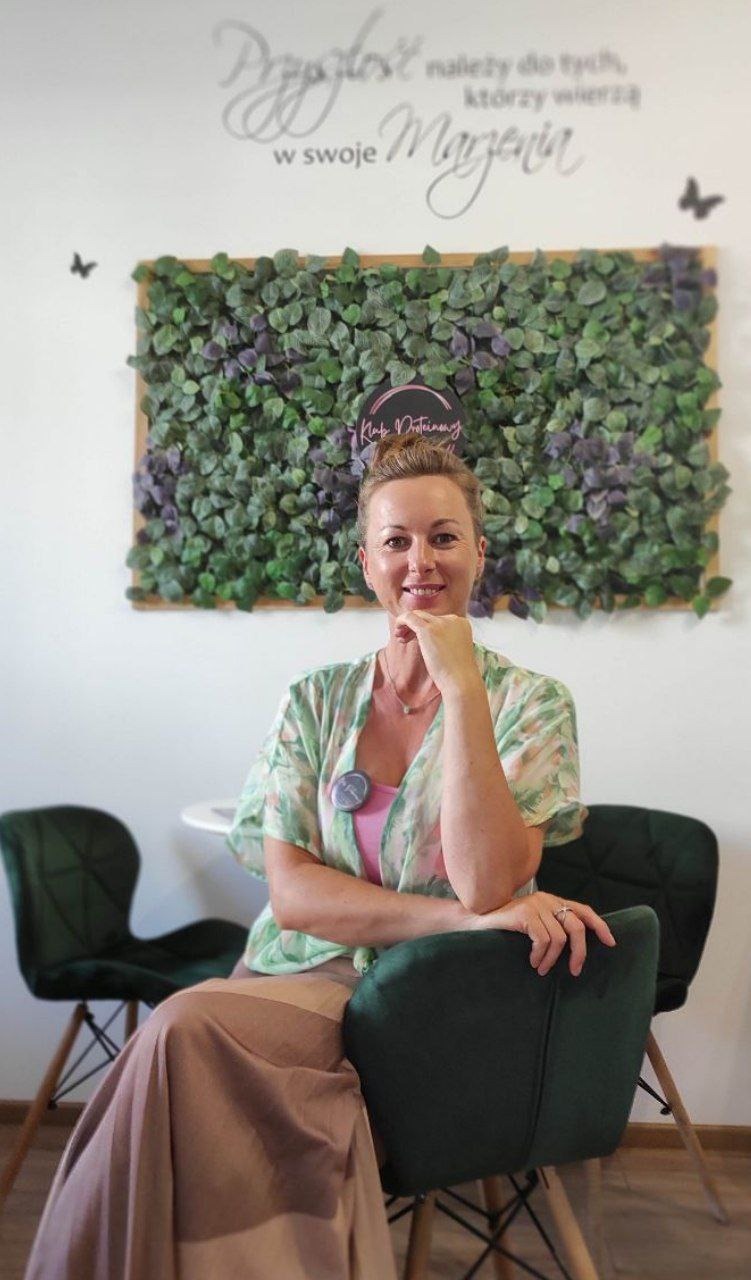 Uśmiechnięta kobieta siedzi w biurze na tle ściany z napisem motywacyjnym i zielonej dekoracji z liści, prezentująca usługi z zakresu zdrowego odżywiania.