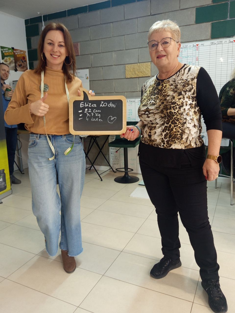 Dwie kobiety prezentują tabliczkę z napisem 'Eliza 20 dni -2,2 cm -3,7 kg - 4 lata' oraz miarkę krawiecką, uśmiechają się, w tle ściana z cegieł i tablica z zapiskami.