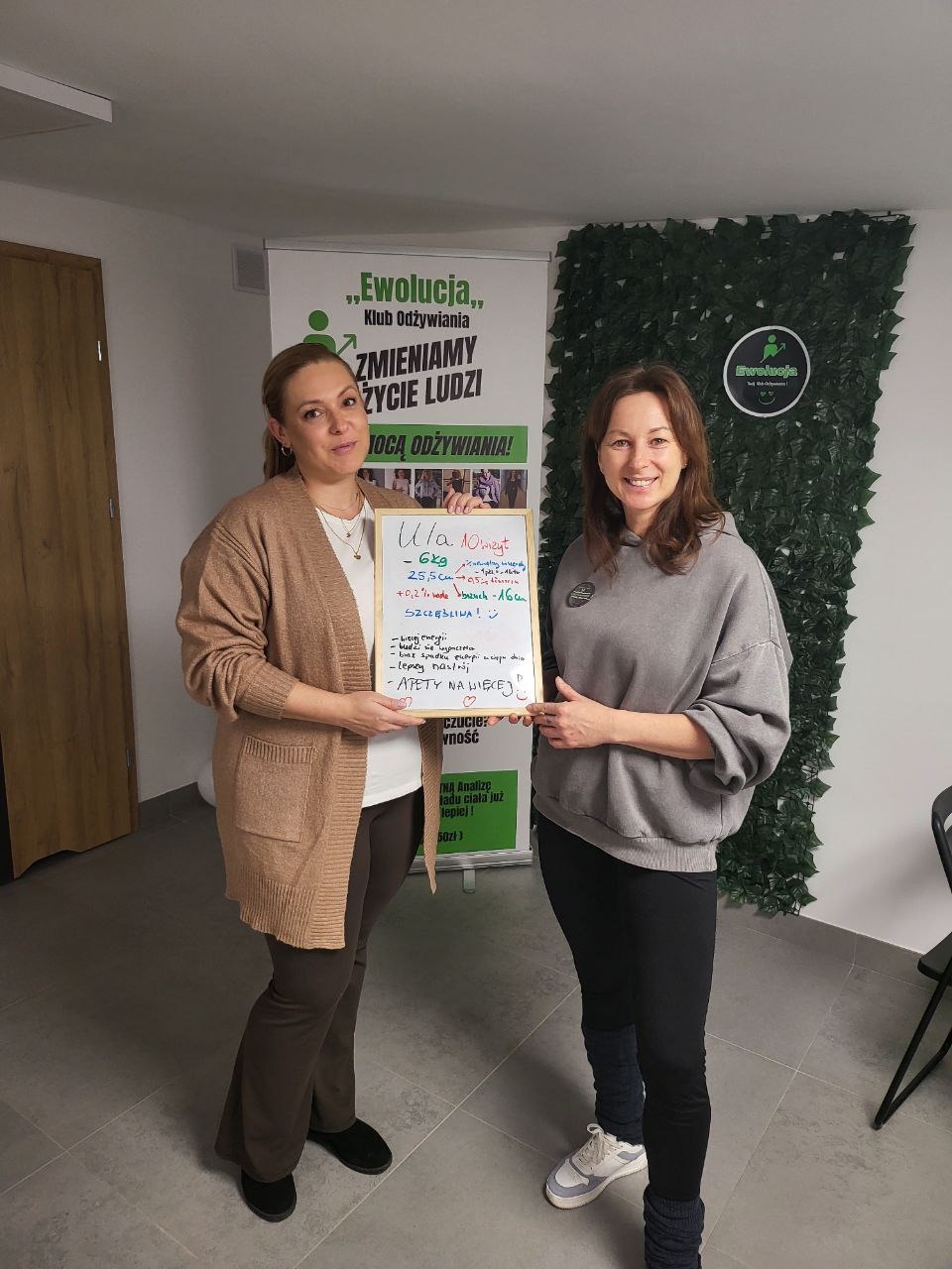 Dwie kobiety prezentują tablicę z wynikami pomiarów ciała, uśmiechnięte, w tle logo 'Ewolucja Klub Odżywiania' i dekoracja z zielonych liści.