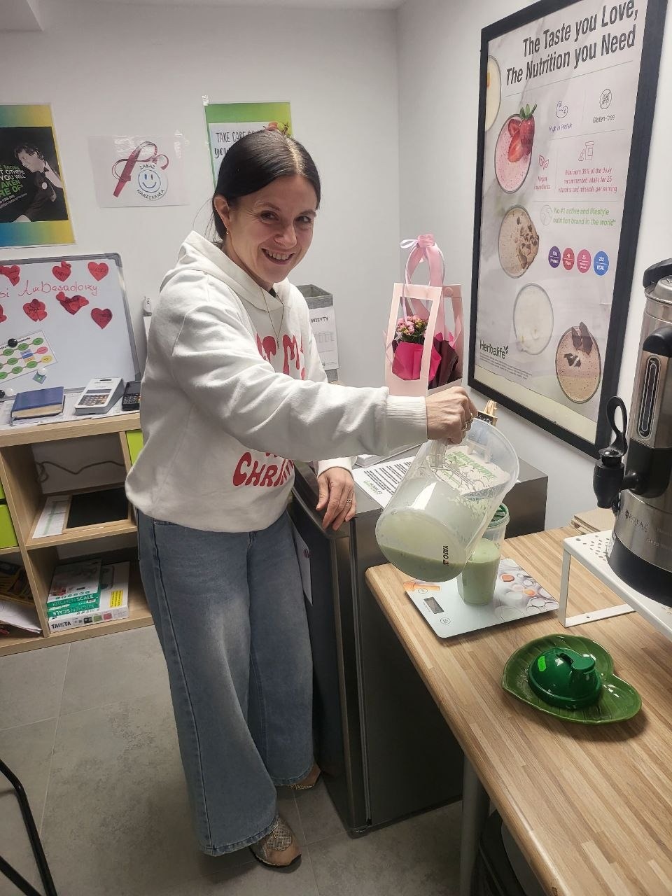 Uśmiechnięta kobieta nalewa zielony koktajl z miarki do kubka na wadze kuchennej. W tle plakat Herbalife i dekoracje.