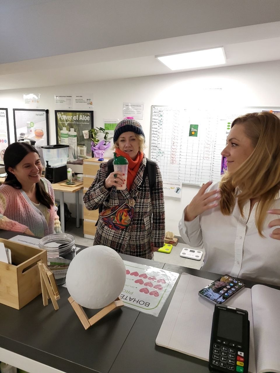 Trzy kobiety w jasnym wnętrzu, jedna trzyma koktajl, na stole waga, terminal płatniczy i materiały informacyjne. Rozmowa o zdrowym odżywianiu lub suplementacji.