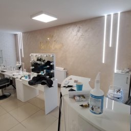 Remonty I wykonczenia wnetrz - Wnętrze salonu kosmetycznego z białymi meblami, dekoracyjną ścianą z napisem 'Bella Ciao' i oświetleniem LED, widoczne akcesoria kosmetyczne i środki czystości.