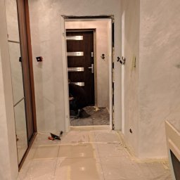 Remonty I wykonczenia wnetrz - Korytarz w trakcie prac: ściany pokryte dekoracyjną gładzią w odcieniach bieli i szarości, zabezpieczona podłoga z jasnych płytek, widoczne narzędzia i sylwetka osoby pracującej przy drzwiach...