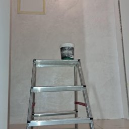 Remonty I wykonczenia wnetrz - Ściana pokryta dekoracyjną farbą Ottocento z efektem aksamitu, widoczna drabina aluminiowa z puszką farby na górze oraz klapa rewizyjna w ścianie.