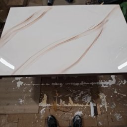 Remonty I wykonczenia wnetrz - Błyszcząca płyta z efektem marmuru na podłodze zabezpieczonej kartonem, widoczne ślady farby, w tle worki z zaprawą i narzędzia.